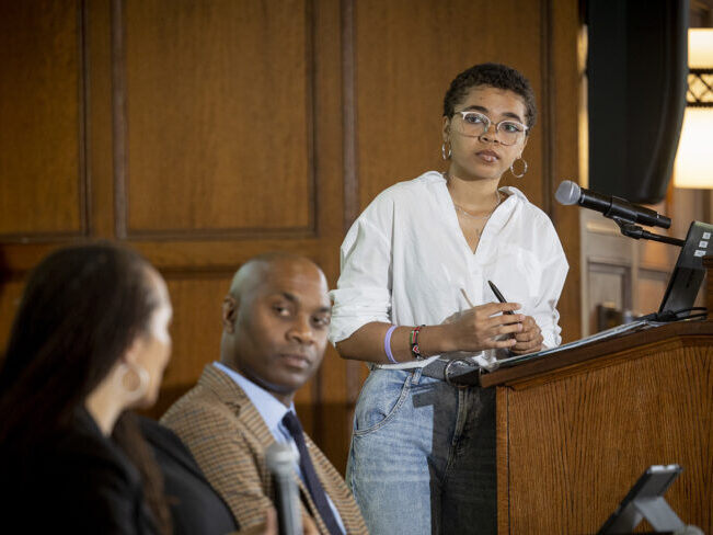 A black student stands at a podium facilitating a panel during the Juneteenth Symposium