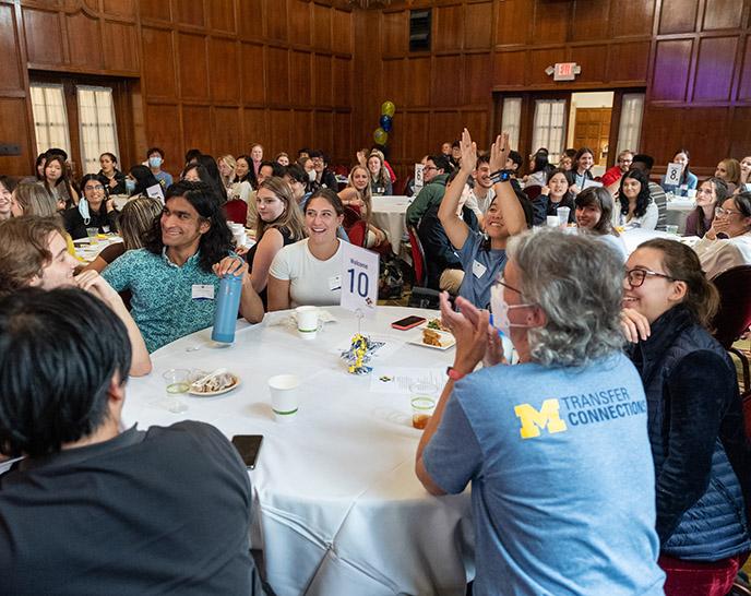 A room full of students sitting at tables are clapping and smiling