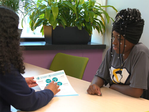 A black student with braids and a SuccessConnects t-shirt listens to a coach explain a studying technique