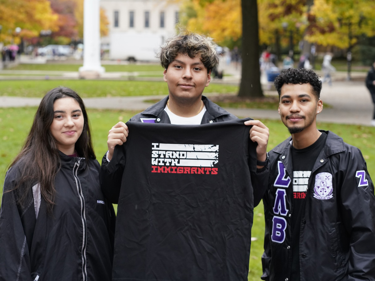 Three students smile and hold up a t-shirt that says 'I Stand with Immigrants'