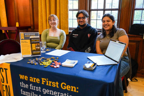 Three students sit at a First-Gen info table during a community dinner