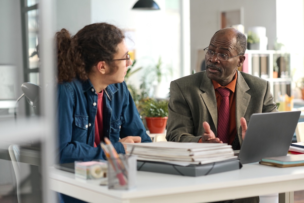 Portrait of senior Black man with young colleague discussing project and using laptop in an office space