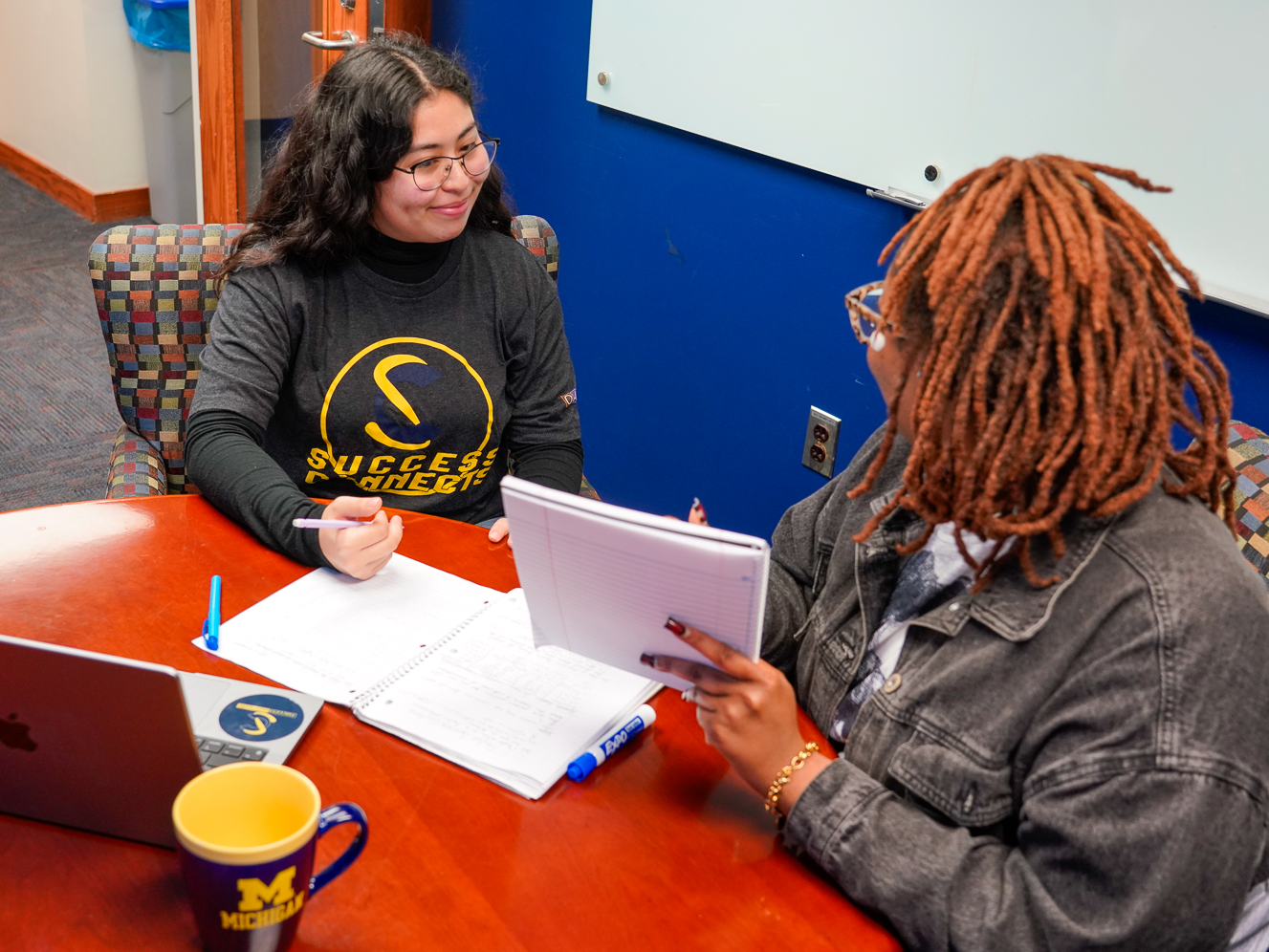 A student is tutored by another student with a laptop and notebooks on the table