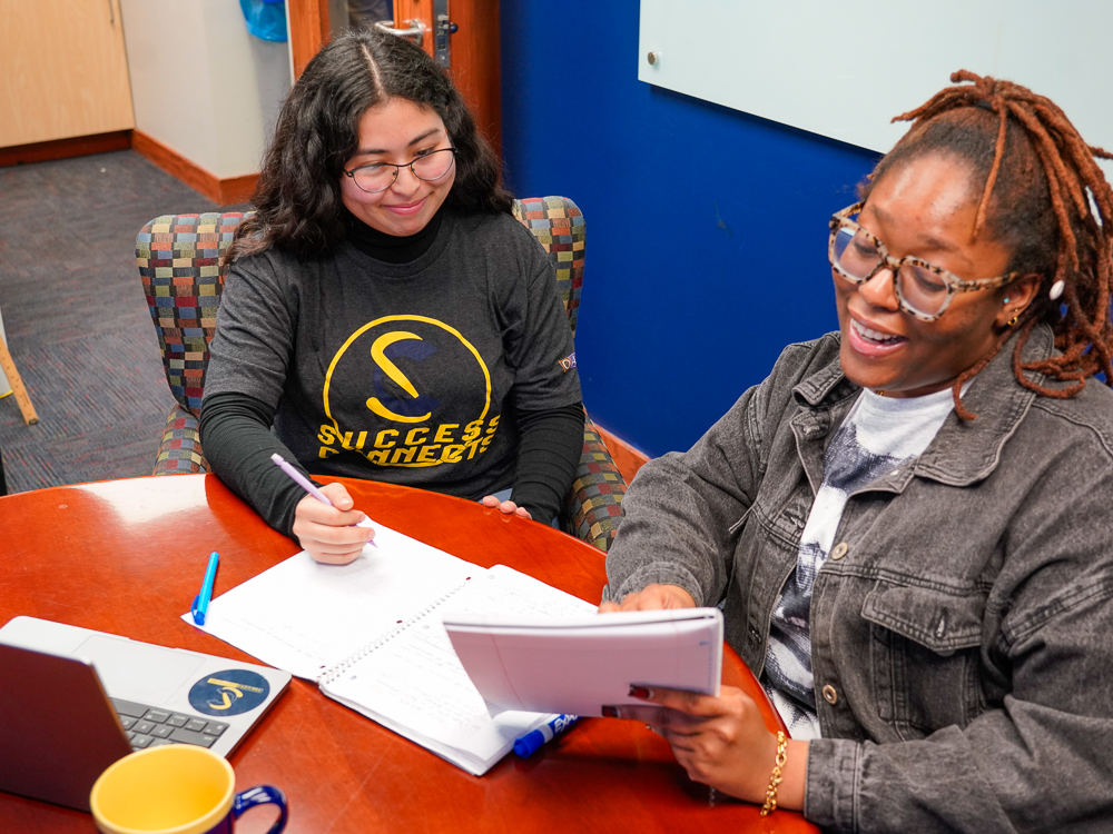 A tutor holds up a notebook and is explaining something to a student wearing a black SuccessConnects t-shirt