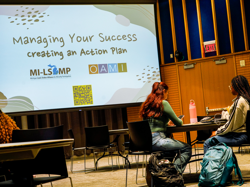 Two students work at a table with a slide presentation in the background that says "Managing Your Success - Creating an Action Plan"
