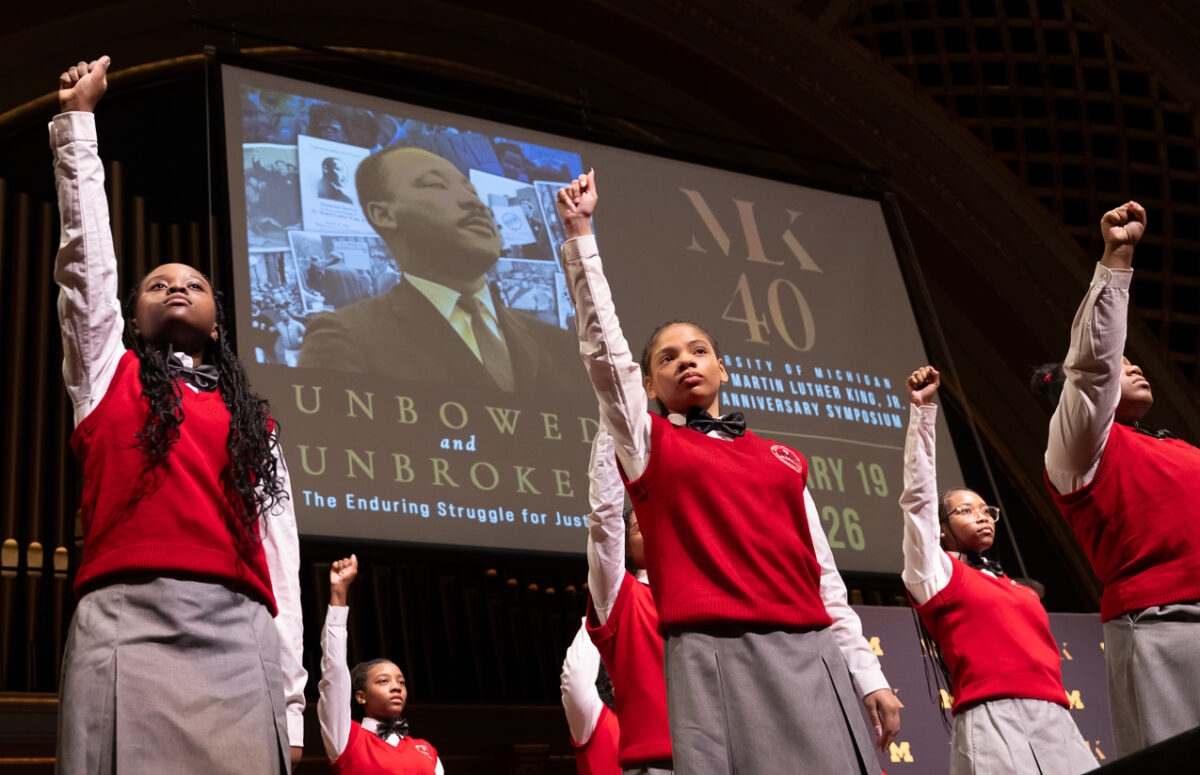 Singers from the Detroit Youth Choir hold up their fists on stage with a graphic of MLK looking up and the event title on the screen behind